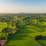 Warm sunset light illuminating the picturesque patchwork quilt landscape of green pasture, agricultural crops, farms and villages below clear blue panoramic skies.