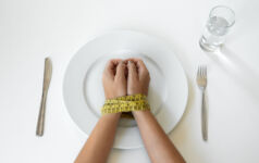 Ozempic vs the ‘sticky’ demand for food Photo of a person's hands bound by a tape measure on a plate, with a knife and fork on either side.