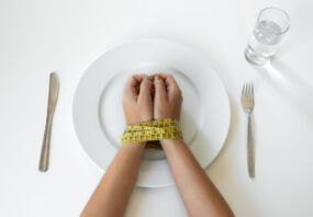 Photo of a person's hands bound by a tape measure on a plate, with a knife and fork on either side.