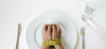 Ozempic vs the ‘sticky’ demand for food Photo of a person's hands bound by a tape measure on a plate, with a knife and fork on either side.
