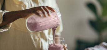 Close-up of hands of an Afro-American woman wearing beige shirt and pouring smoothie into a glass on a kitchen table. There is a part of a blender, a cutting board and a cloth on the desk in front of her. In the corner of a room sits a big plant.